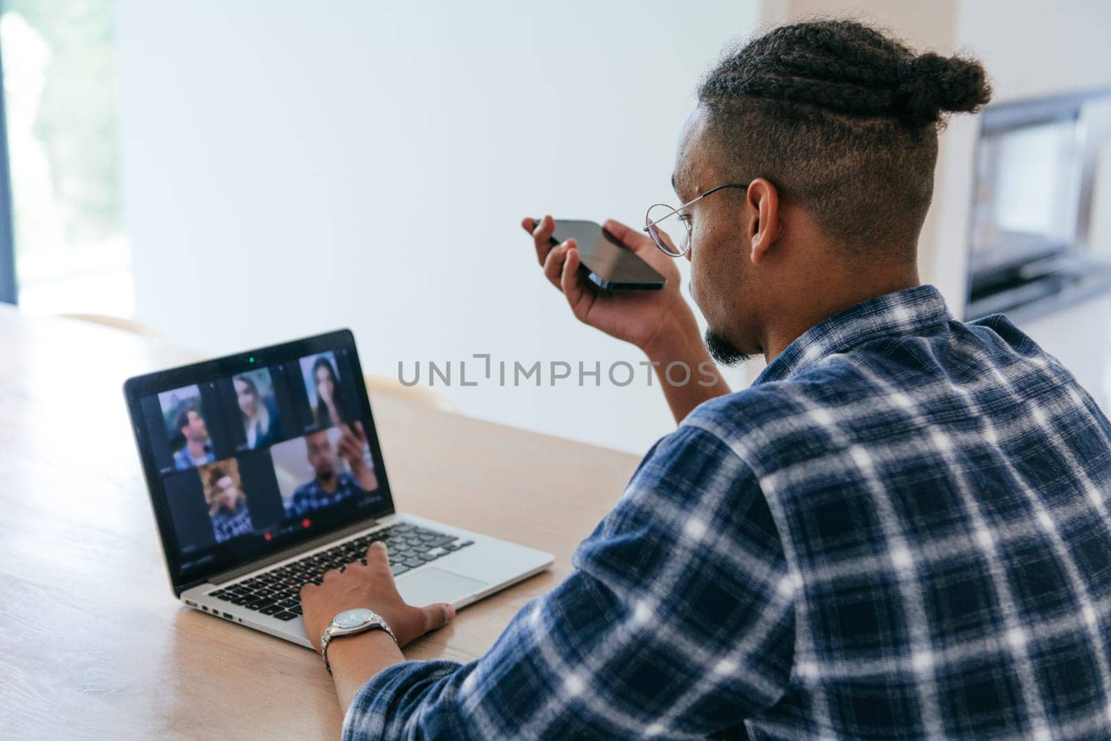 African American businessman in glasses sitting at a table in a modern living room, using a laptop and smartphone for business video chat, conversation with friends and entertainment