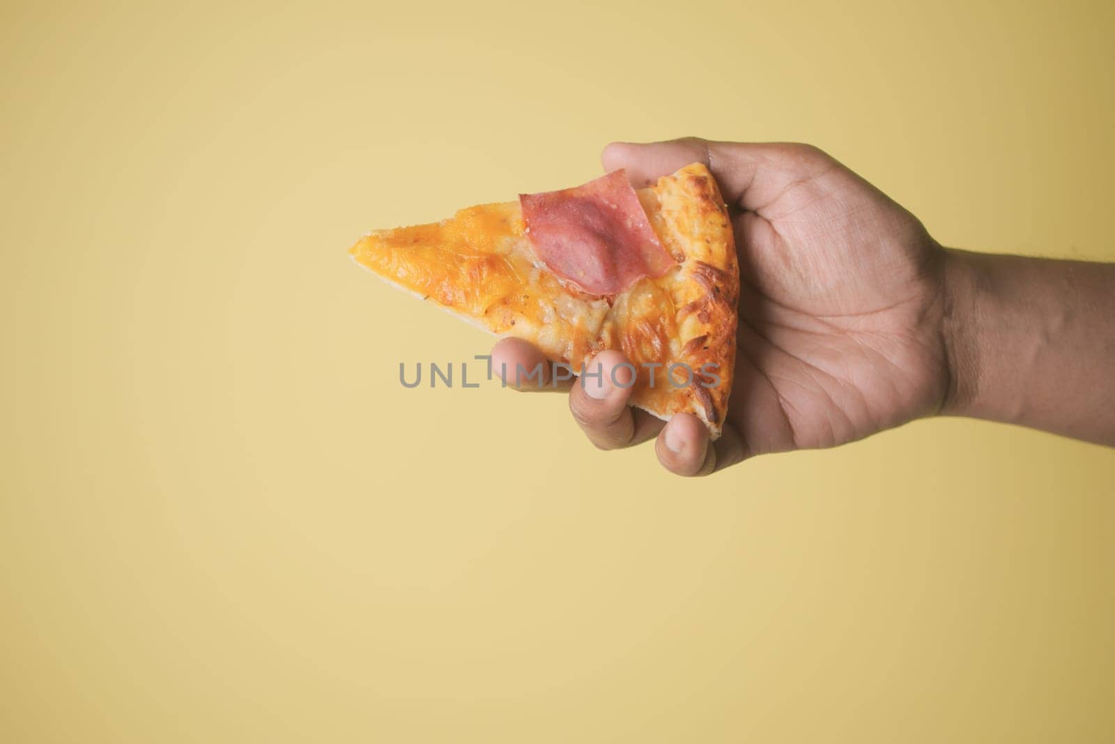 A hand holds a slice of pizza topped with cheese and ham on a yellow background.