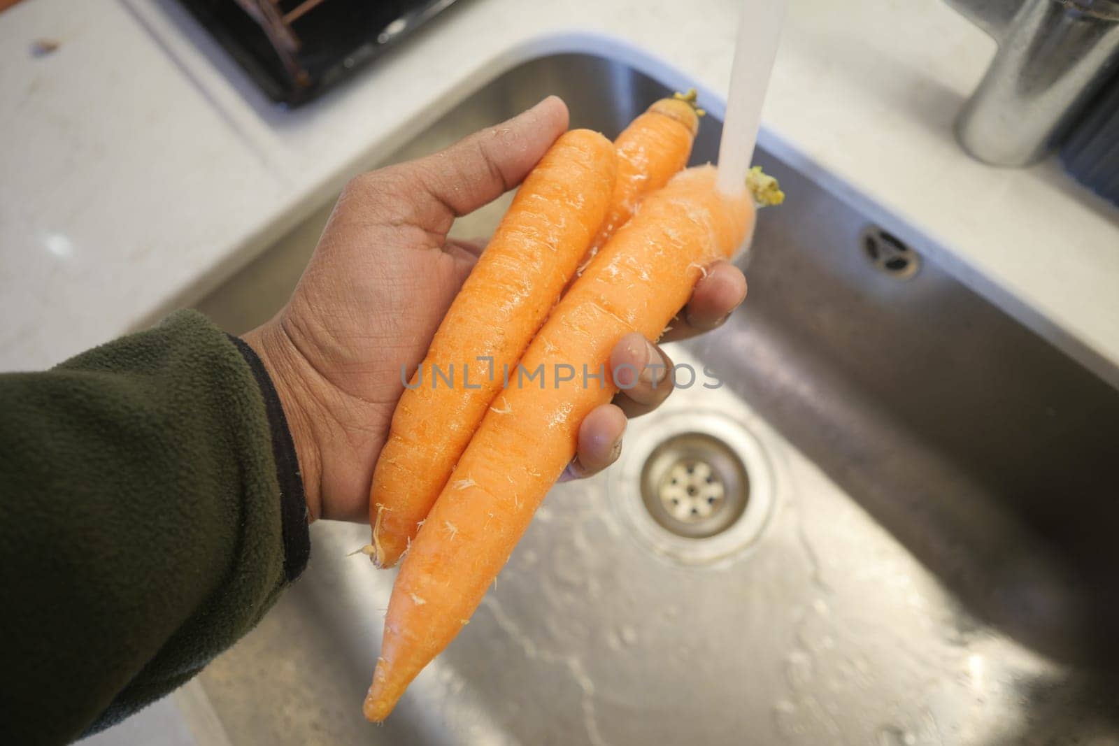 Hand holding clean, orange carrots over a stainless steel sink in kitchen.