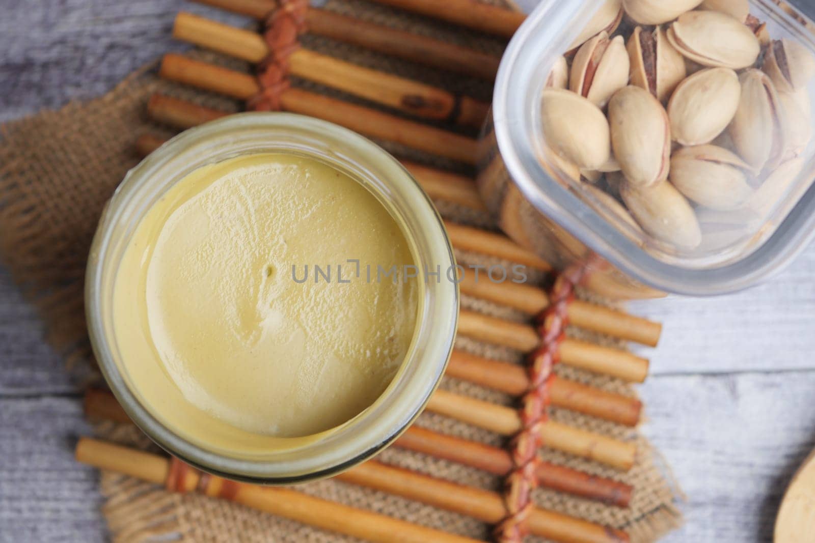 Pistachio spread sits in a clear glass jar beside shelled nuts on a mat.
