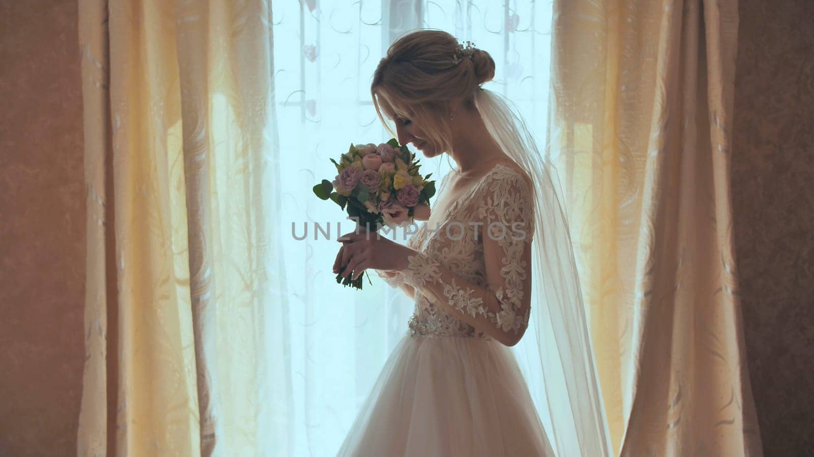 Beautiful blonde bride holds flower bouquet near the window.