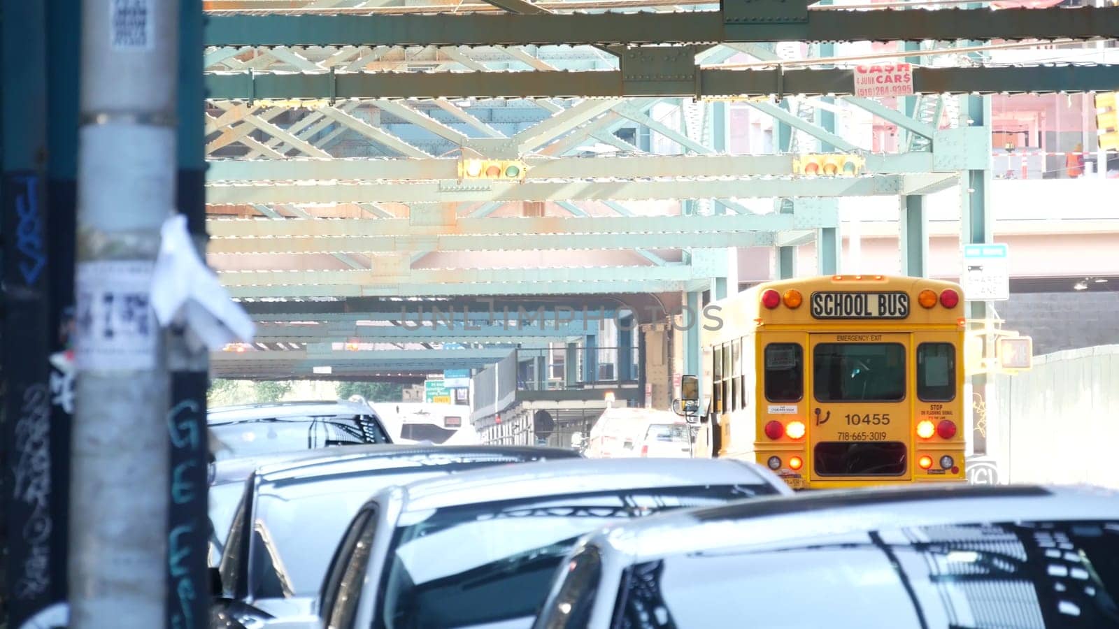 New York City, United States - 31 Aug 2023: Elevated subway above street. Metropolitan bridge over road, Queens. Overhead metro line. Car under railway track. Urban life. Yellow school bus, education.