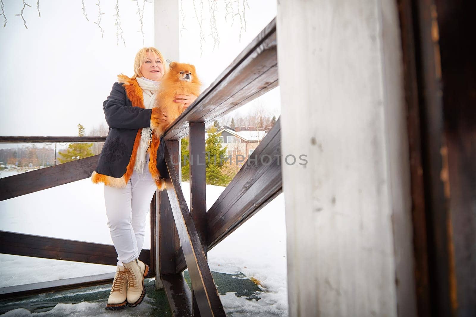 Middle-aged woman stands by a railing with her fluffy Pomeranian, surrounded by snow-covered scenery on a winter day.