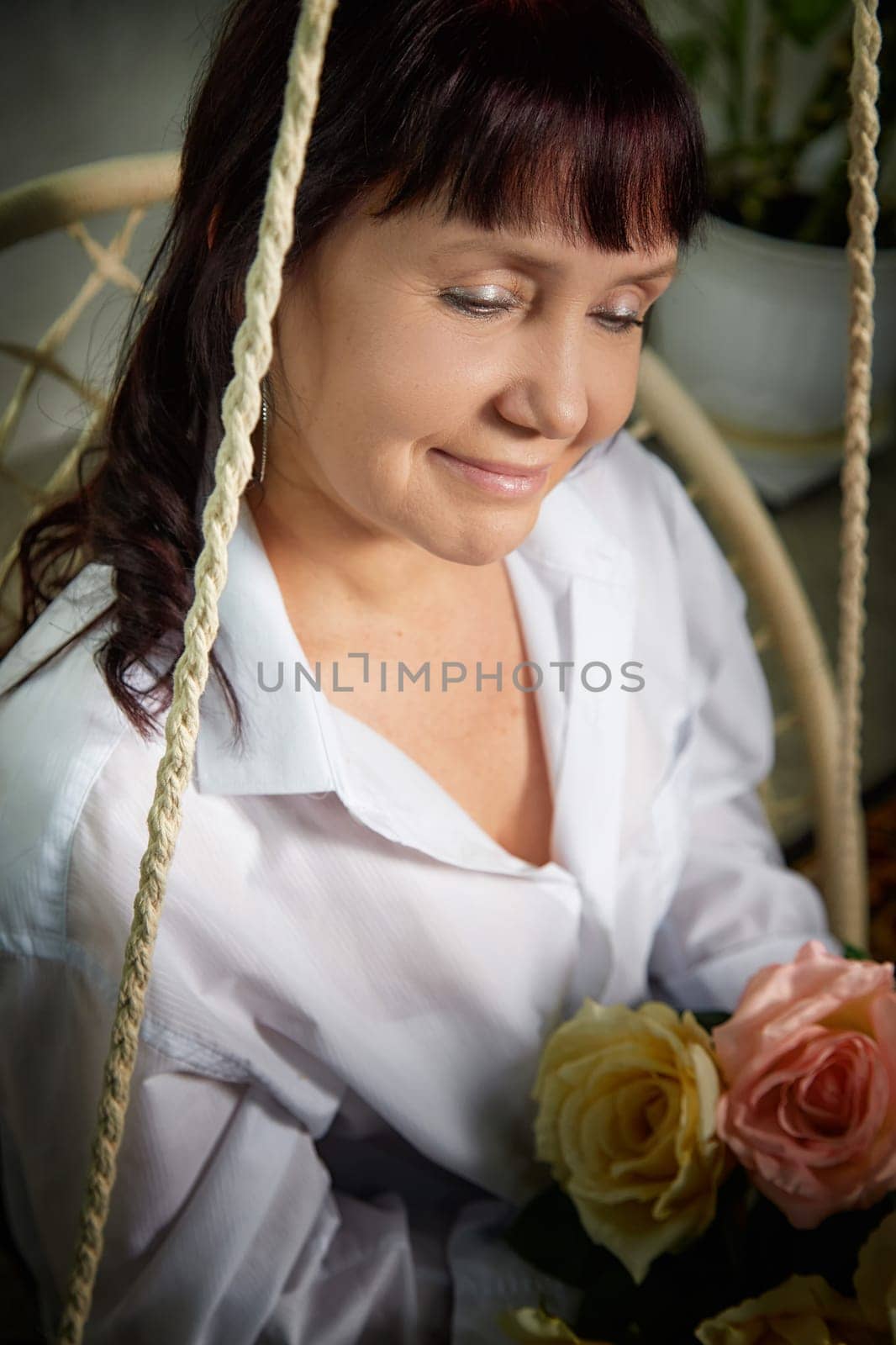 A middle-aged woman sits gracefully on a swing indoors, holding a bouquet of roses while smiling warmly in her stylish apartment.