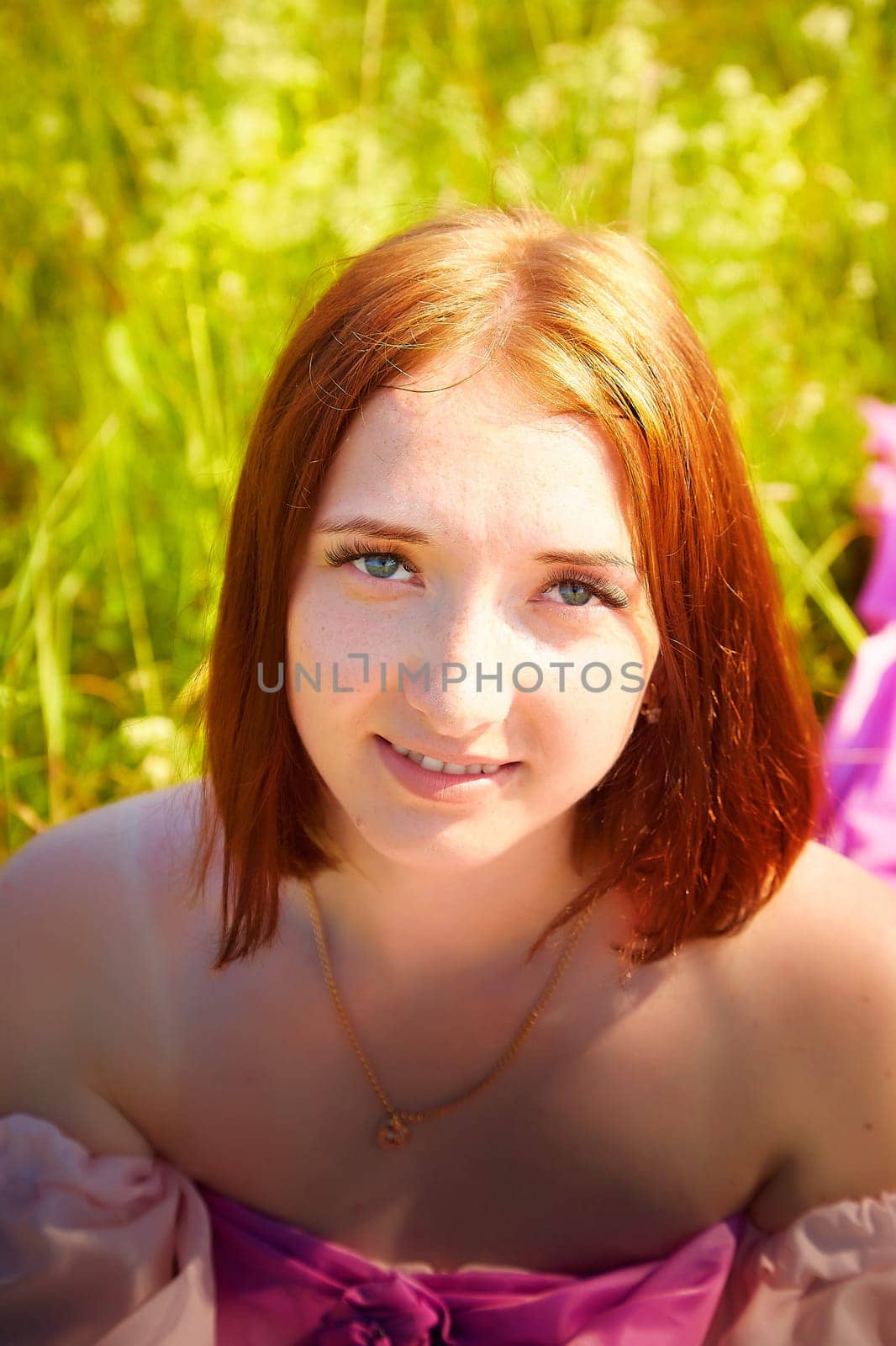 Bright sunlight illuminates a young woman with red hair sitting in a vibrant meadow filled with lush green grass and wildflowers.