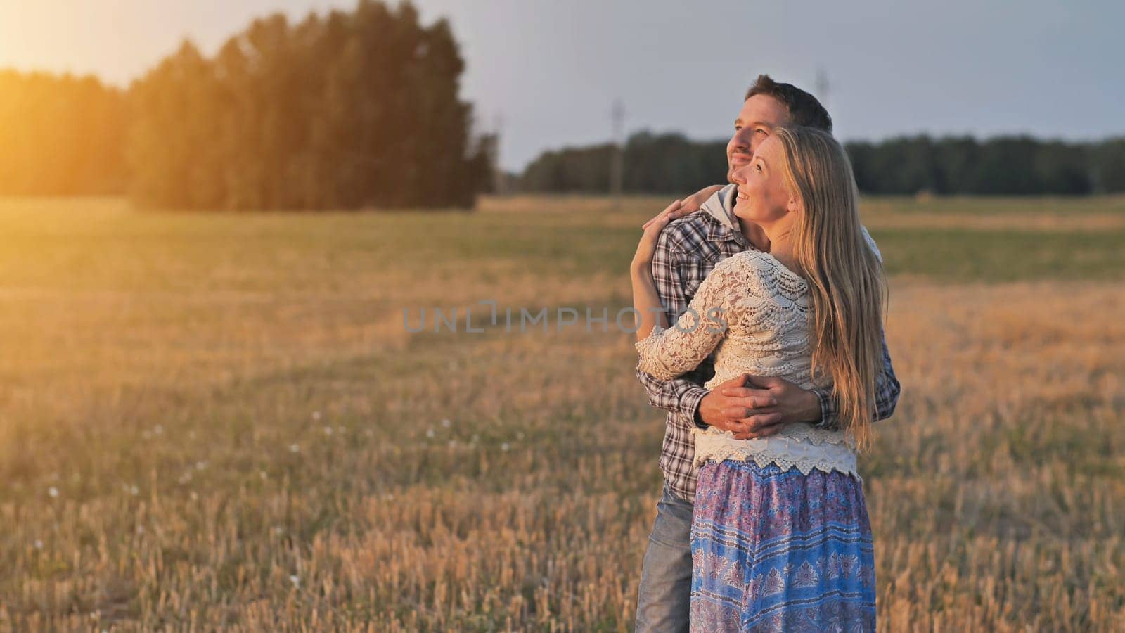 Young husband and wife cuddle at sunset and look towards the sun.