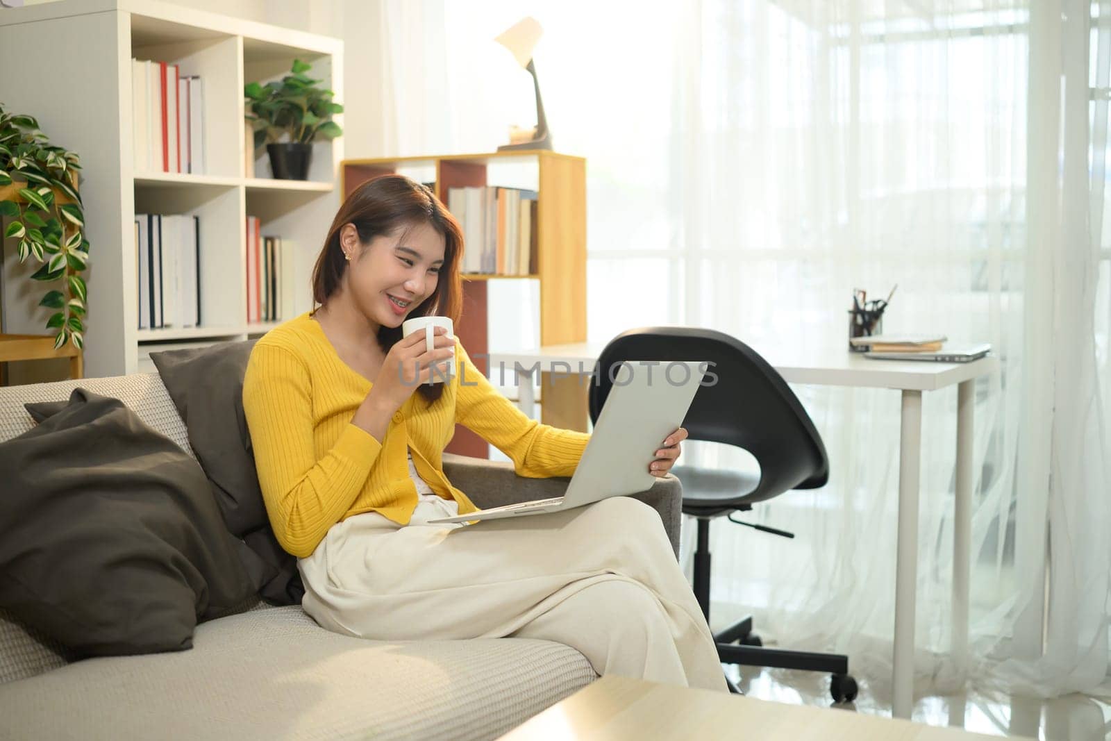 Cheerful asian woman holding a coffee mug and browsing on her laptop in living room.
