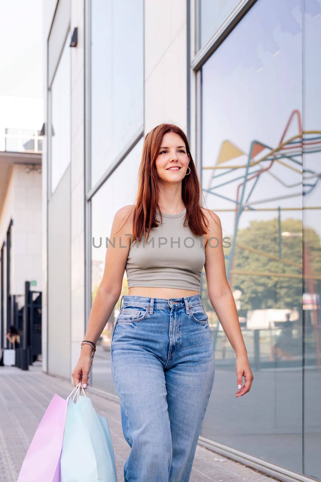 young adult woman in casual clothes walking down a sidewalk near of a mall carrying shopping bags, concept of youth and urban lifestyle, copyspace for text