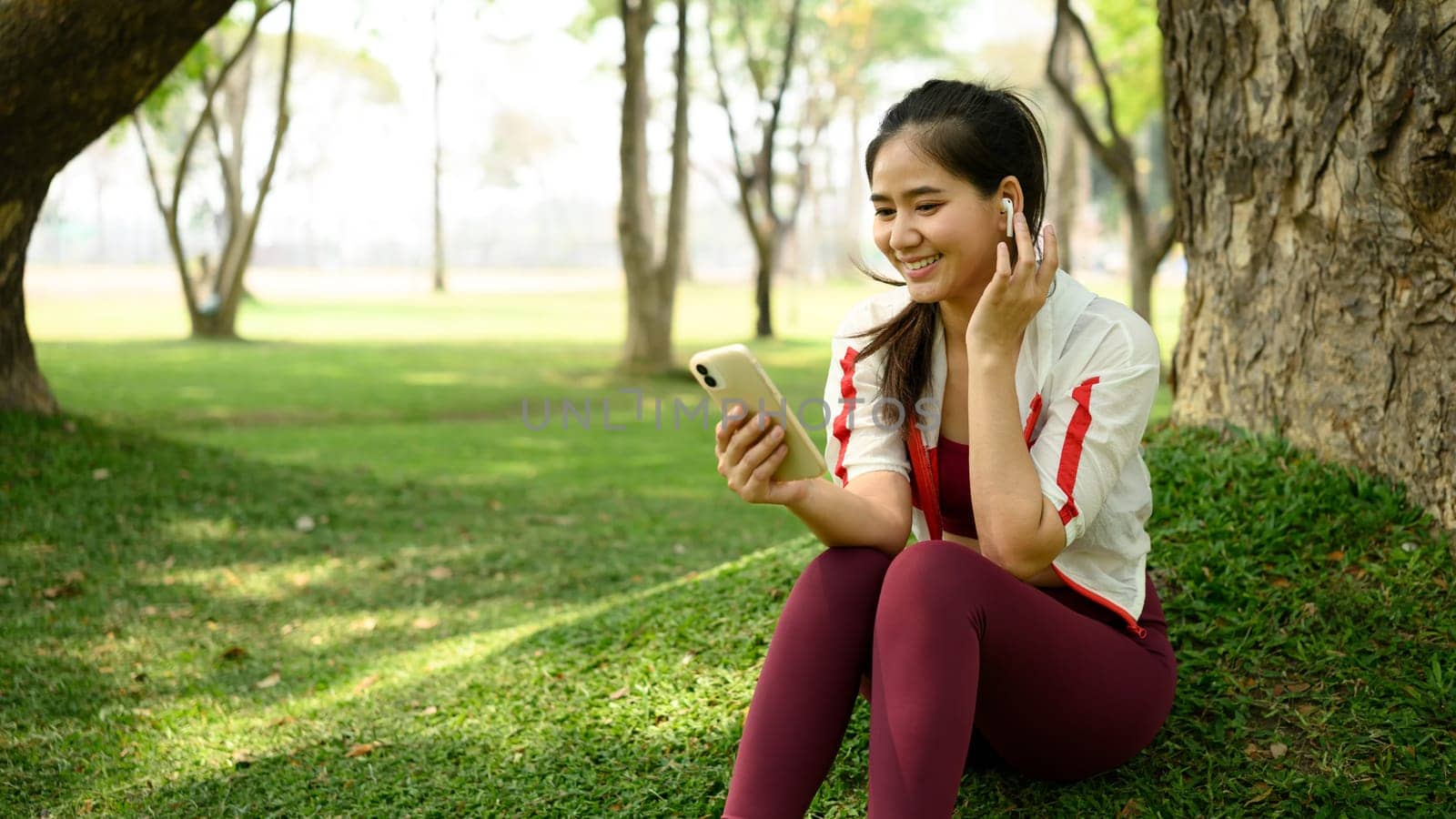 Smiling young woman sitting under a tree in a park, using her phone and wireless earbuds.