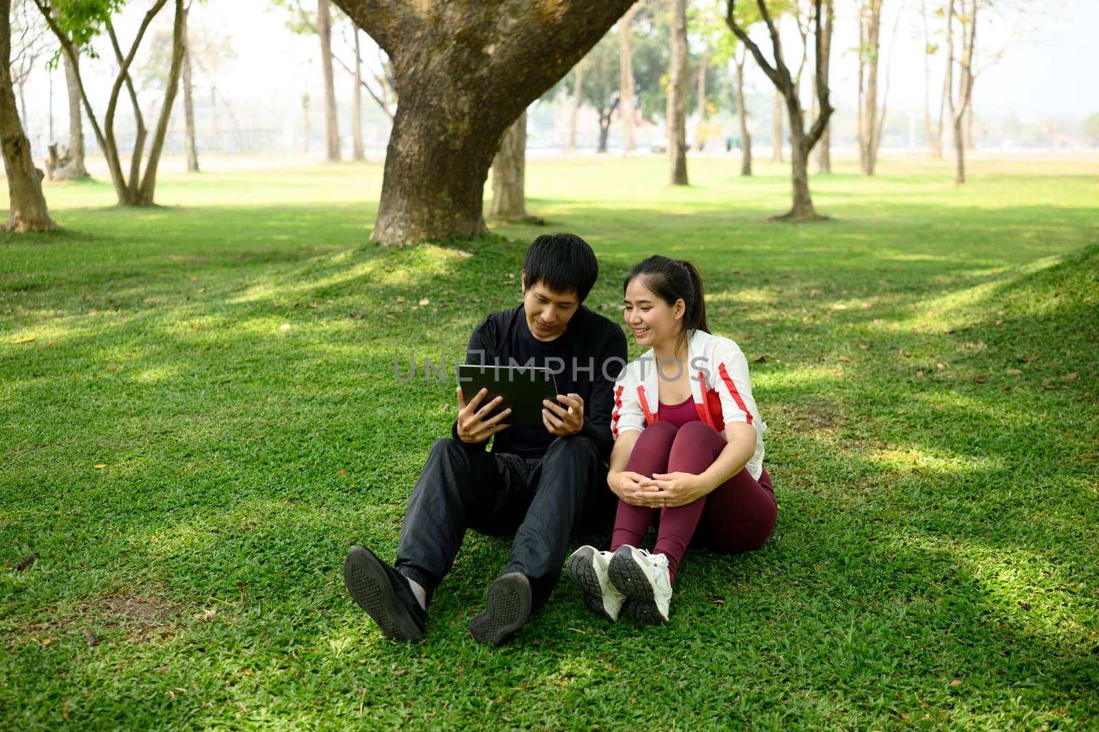 Asian couple checking workout plan on digital tablet, sitting on yoga mats in a sunny park.