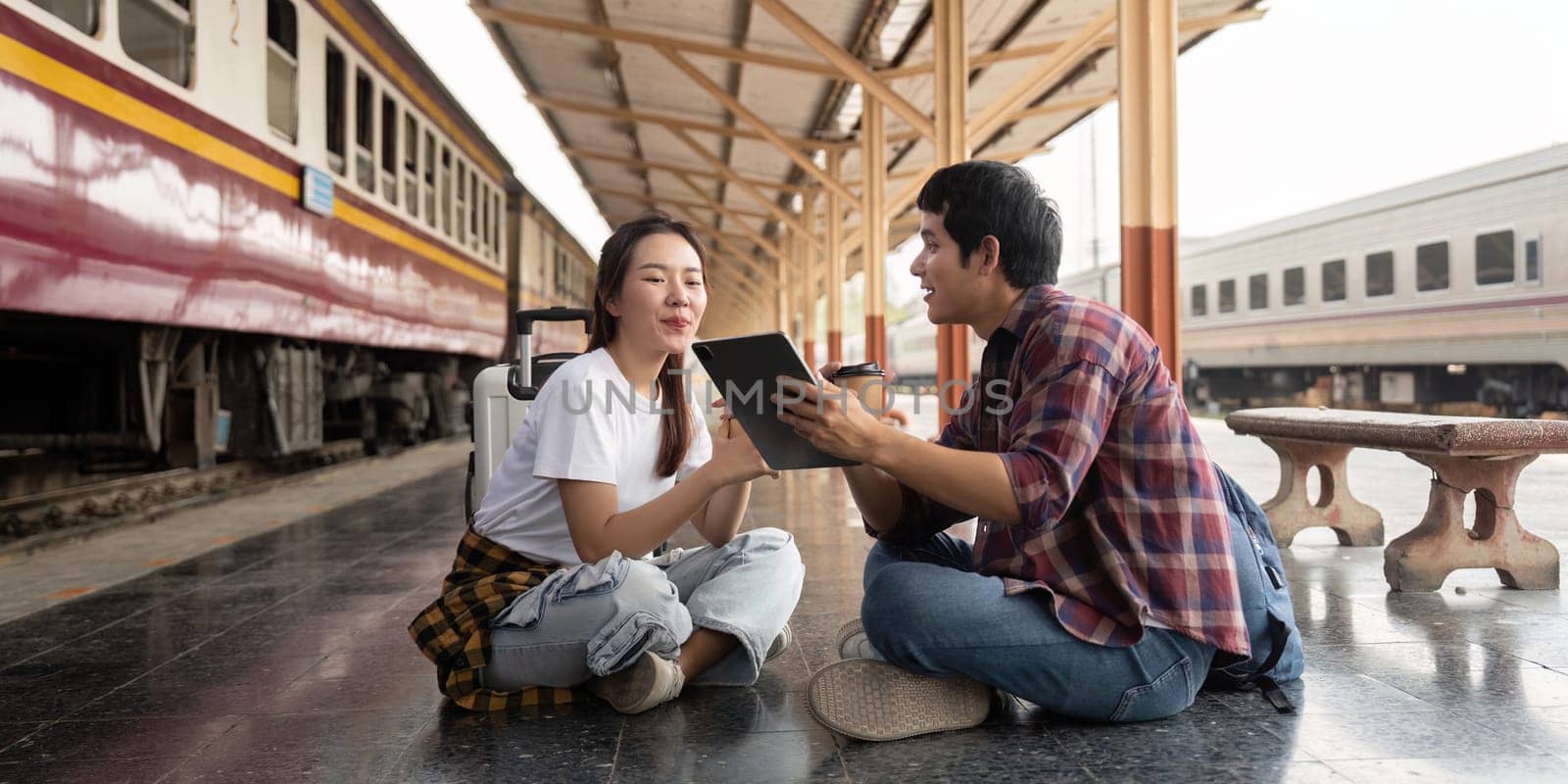 At a bustling train station, a couple sits on the ground, engaged in a discussion about their upcoming travels. The excitement of their journey ahead shines through.
