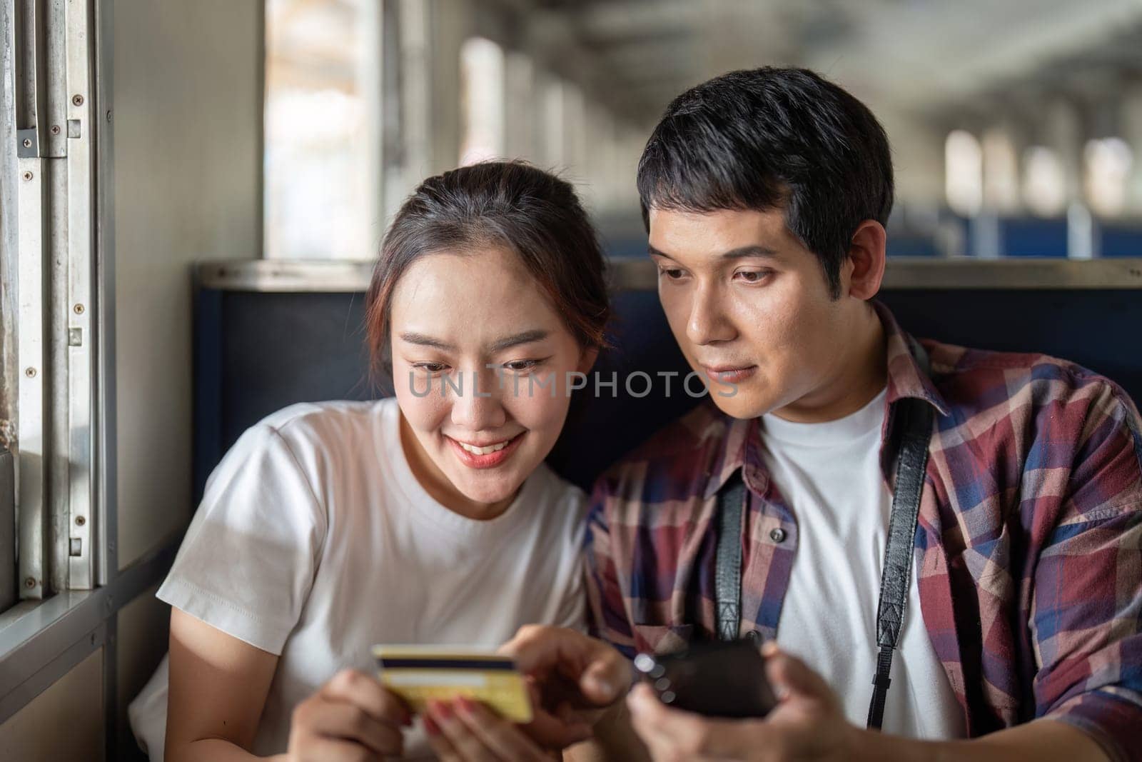The couple shares a lighthearted moment on the train, using their devices and smiling at each other, representing modern travel companionship.