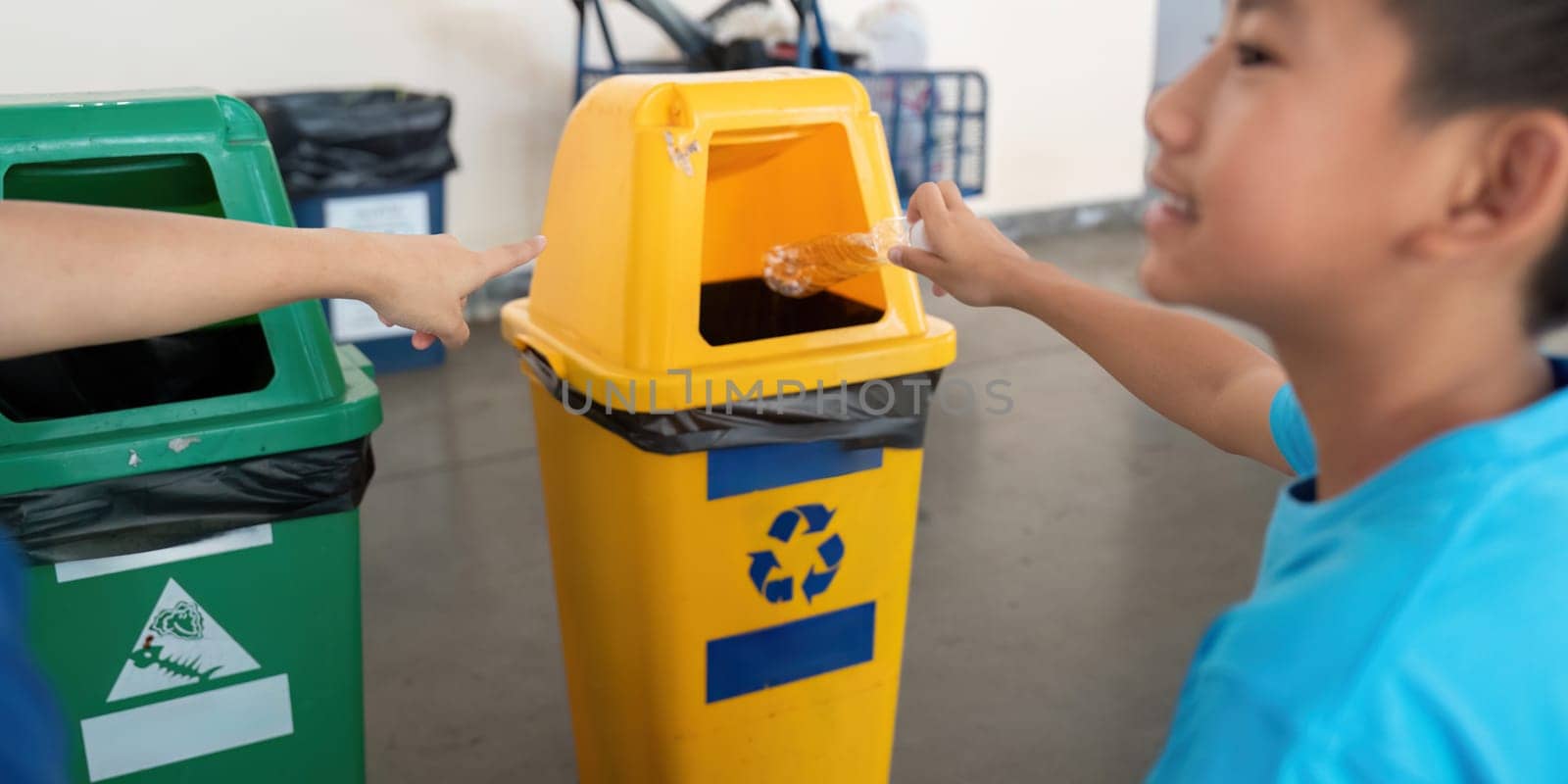 A young boy enthusiastically discards plastic waste in a recycling bin, guided by a supportive instructor in a sustainability initiative. The moment emphasizes environmental education.
