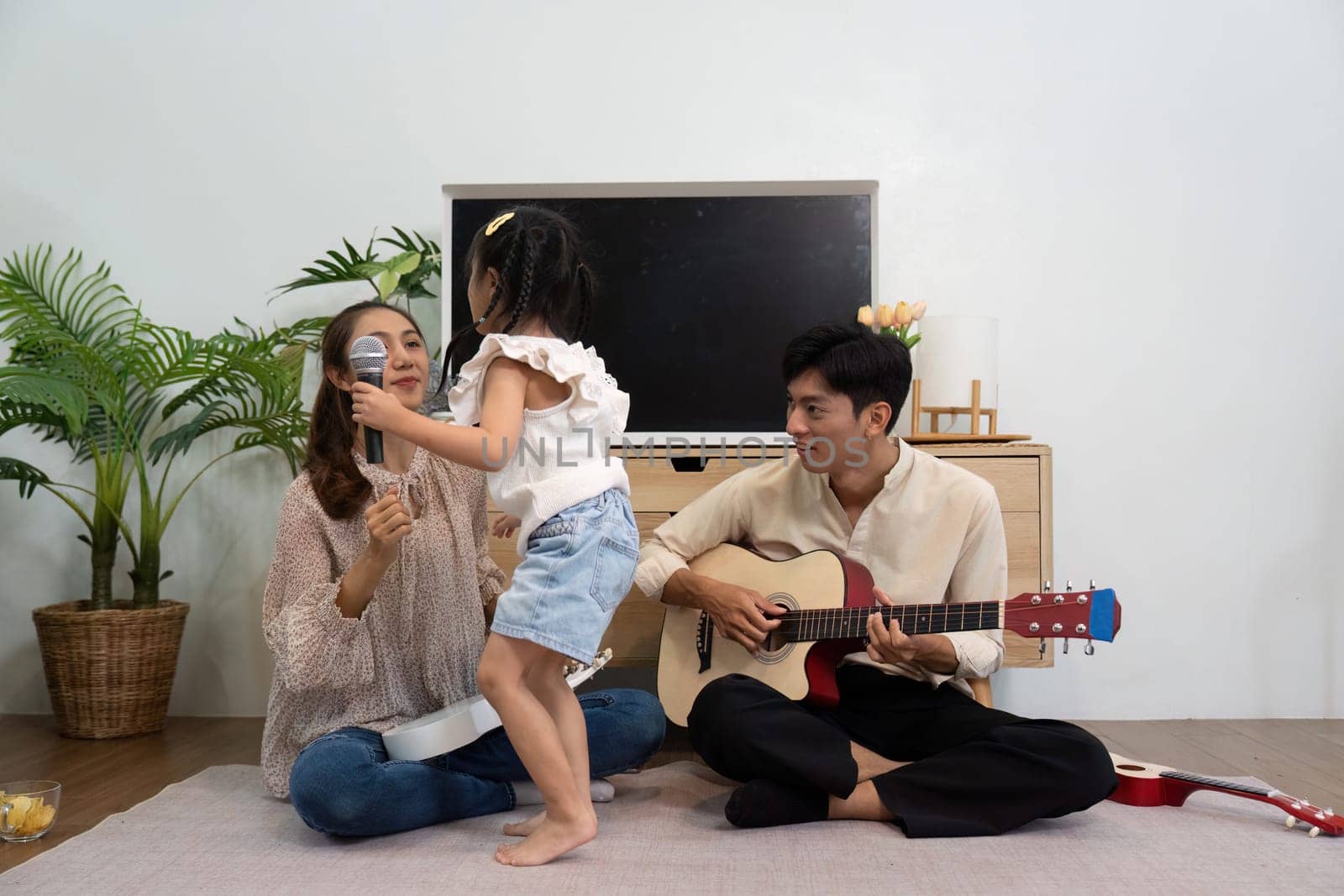 A lively scene as a little girl switches between a microphone and a ukulele, surrounded by her loving parents, radiating fun and creativity.