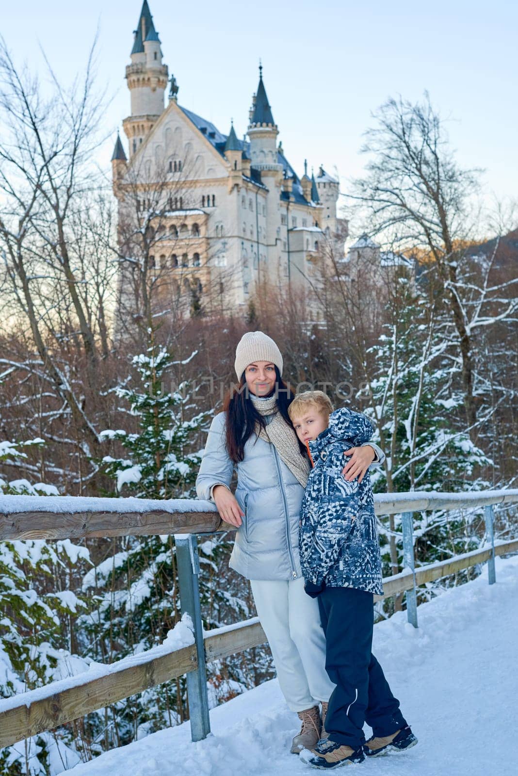 Enchanting winter lifestyle portrait capturing tender moment between mother and young boy exploring magical snow covered castle grounds. Elegantly dressed woman in contemporary winter outerwear holds hands with adorable child while standing on rustic wooden bridge surrounded by pristine winter scenery. Majestic historic castle with ornate towers provides stunning architectural backdrop against crystal clear blue sky. Natural authentic moment showcases family connection, winter tourism, fairytale atmosphere and European cultural heritage perfect for travel, tourism, parenting, and winter lifestyle marketing. Mother and Child Exploring Snowy Winter Castle Wonderland on Wooden Bridge Against Blue Sky with Historic Architecture and Snow Covered Landscape