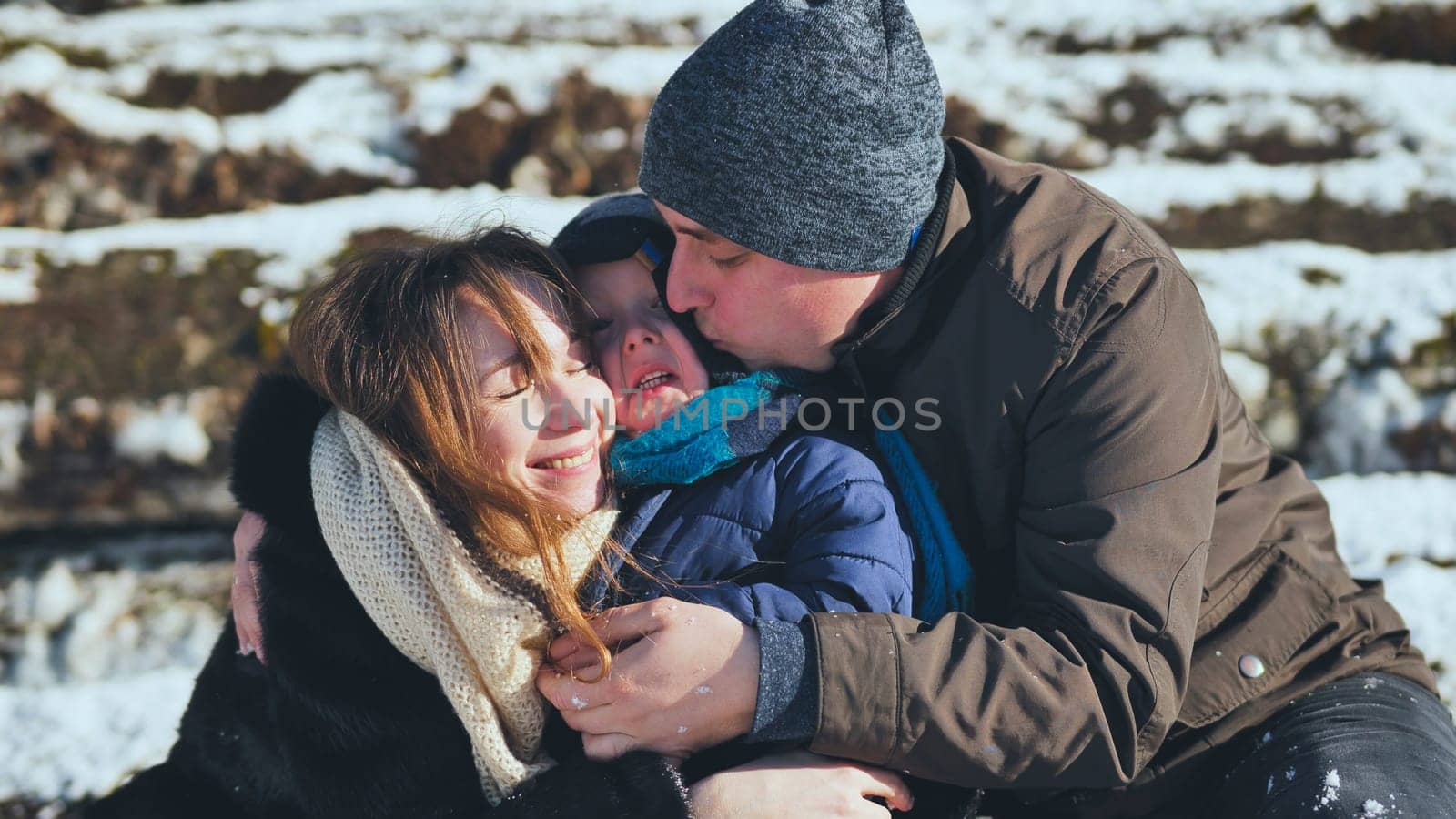Parents hugging and kissing their joyful son on a chilly winter day, savoring the beauty of a picturesque snowy landscape together