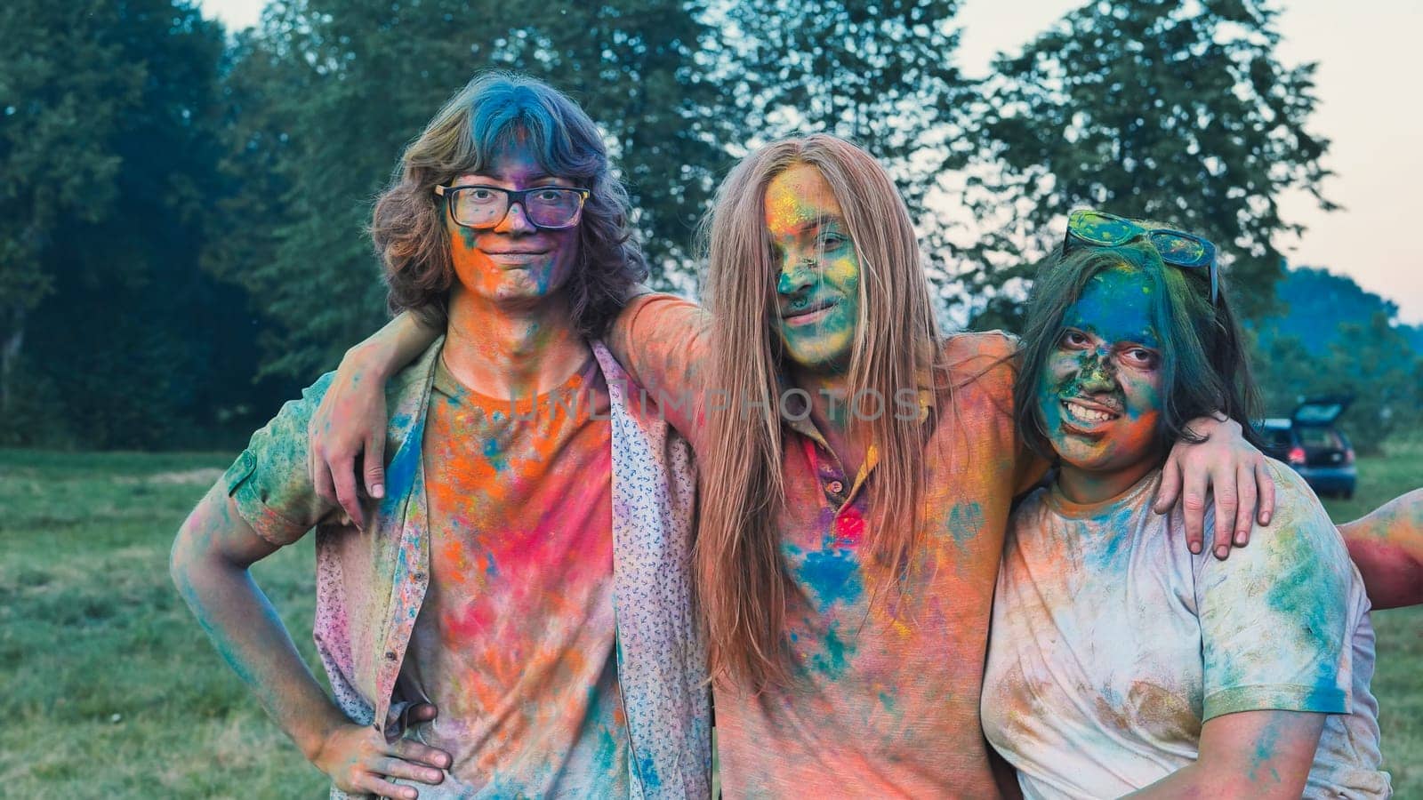 Three school friends are embracing each other, covered in vibrant holi powder, enjoying the colorful festivities