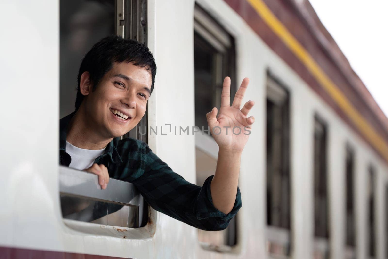 A young man happily waves from the window of a moving train, embracing the excitement of exploration and the thrill of adventure on his travels.