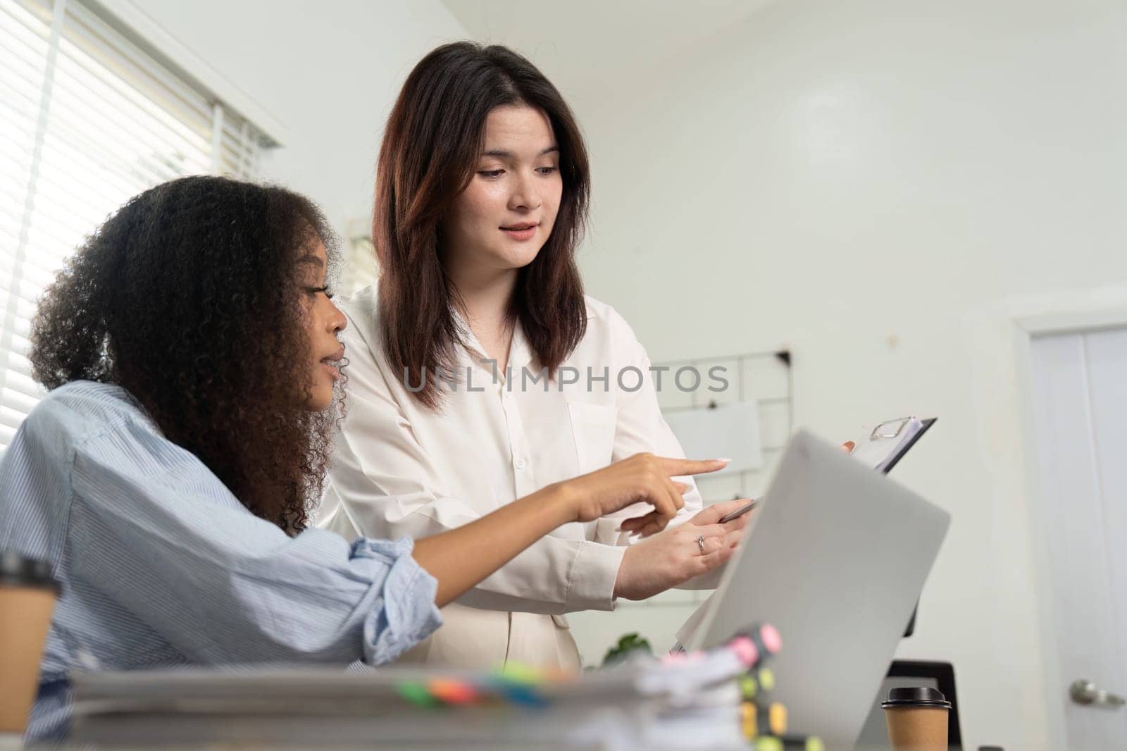 In a vibrant workspace, two women collaborate on solutions while reviewing information displayed on a laptop, emphasizing teamwork and innovation.