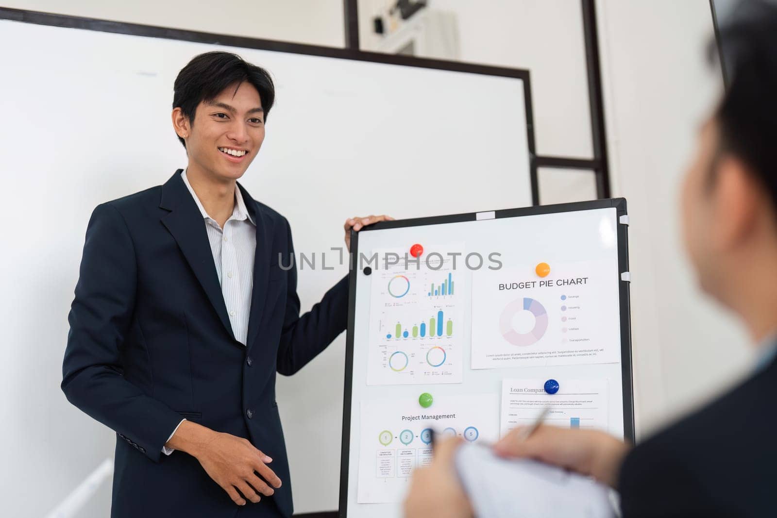 A confident male presenter explains a budget pie chart, engaging with team members who are attentively listening and taking notes. The setting emphasizes teamwork and effective communication.