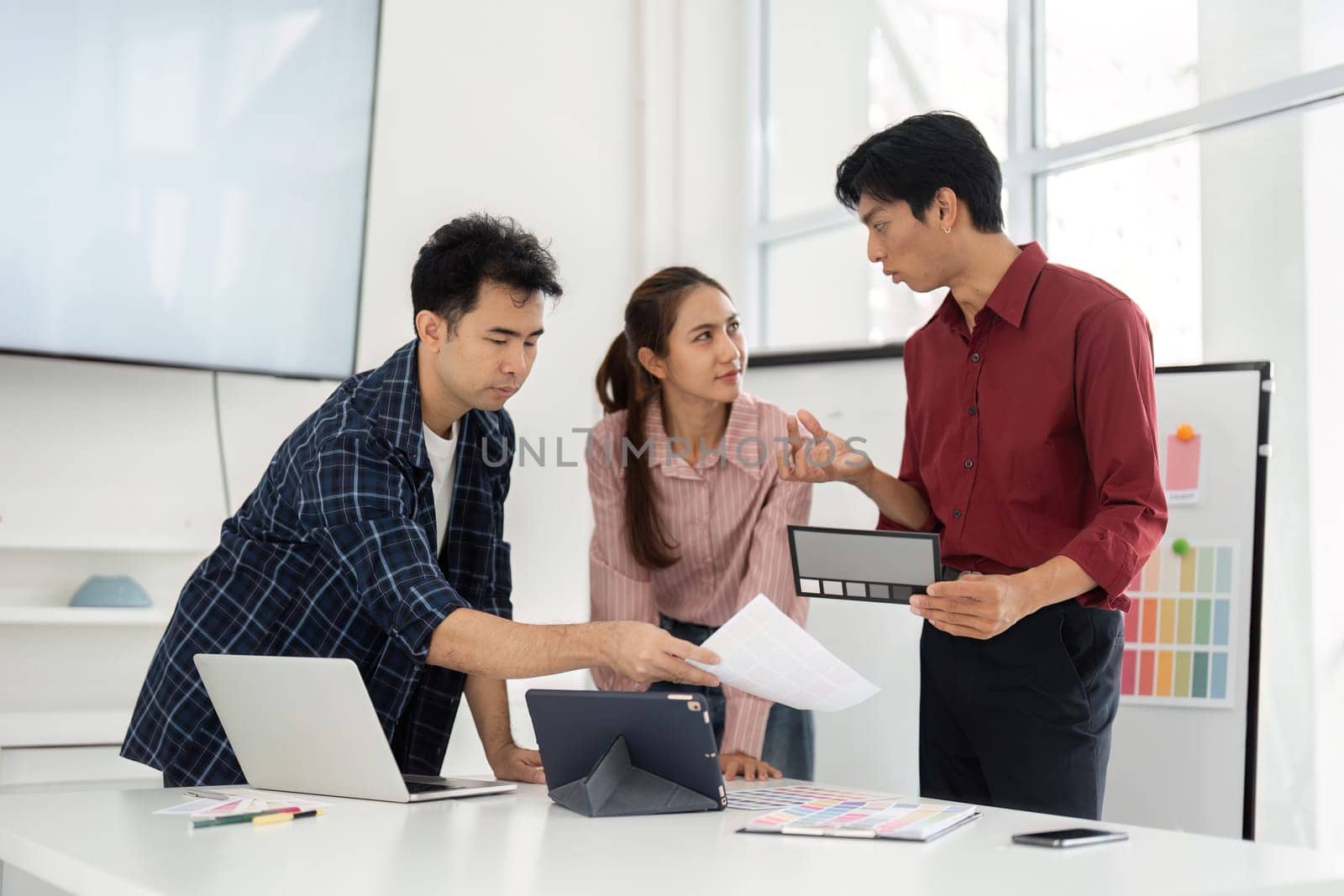 Three team members discuss innovative design ideas while analyzing color samples on their tablets and papers in a bright workspace, exemplifying teamwork and creativity.