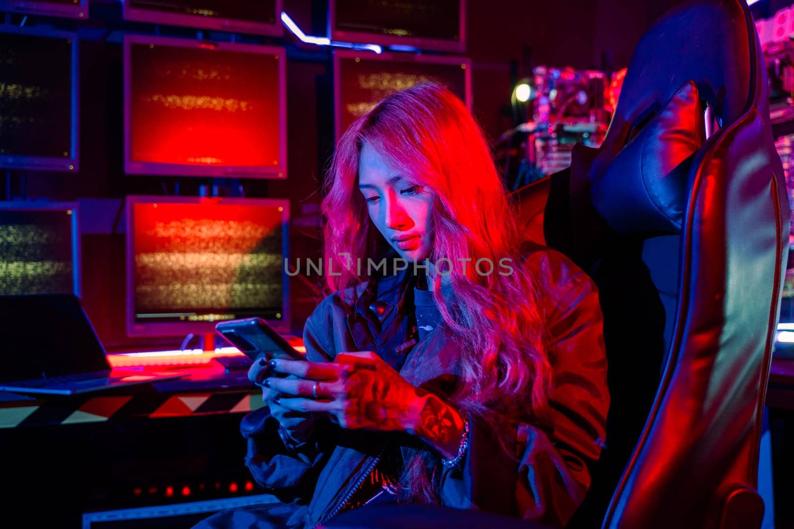 Asian woman in front of a computer monitor display using mobile phone, neon light, Hacker wear black jacket at system control room, Security guard working on multiple monitors together smartphone