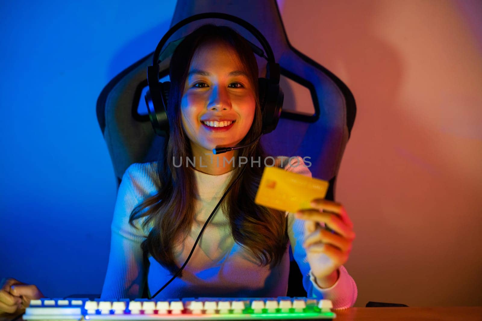 Asian gamer playing online video game and holding a credit card for payment game online, young woman in gaming headphones using computer for playing games at home, broadcast streaming live, Front view