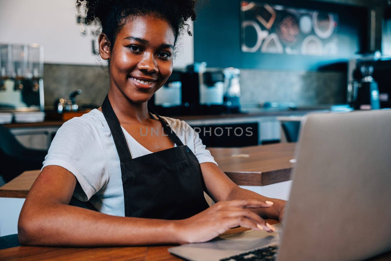 Portrait of a self-employed black woman barista at her coffee shop counter working on laptop. Confident entrepreneur managing finance bookkeeping in her own business.