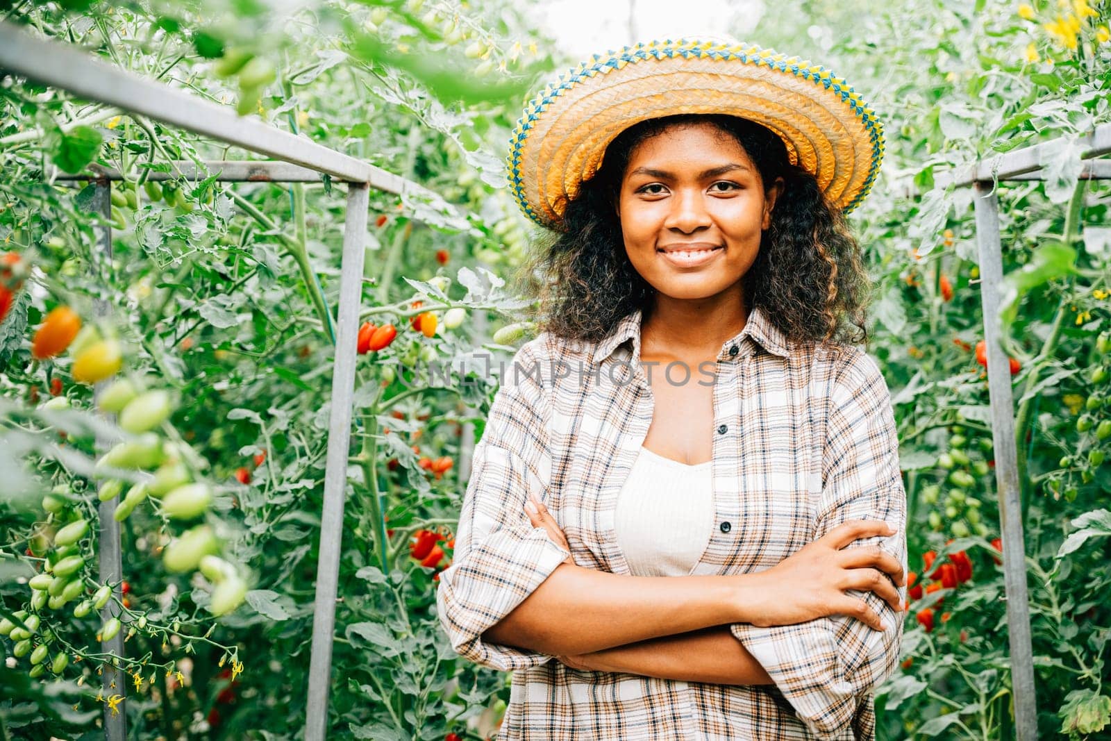 A cheerful woman farmer arms crossed tends to vegetables in a tomato greenhouse. Her smiling portrait reveals dedication and happiness in nurturing growth outdoors.