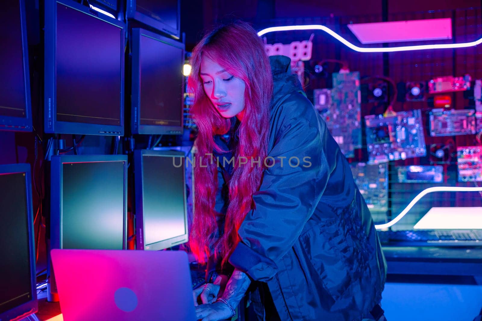 Asian woman in front of a computer monitor display big digital data with laptop computer, neon light, Hacker wear black jacket at system control room, Security guard working on multiple monitors