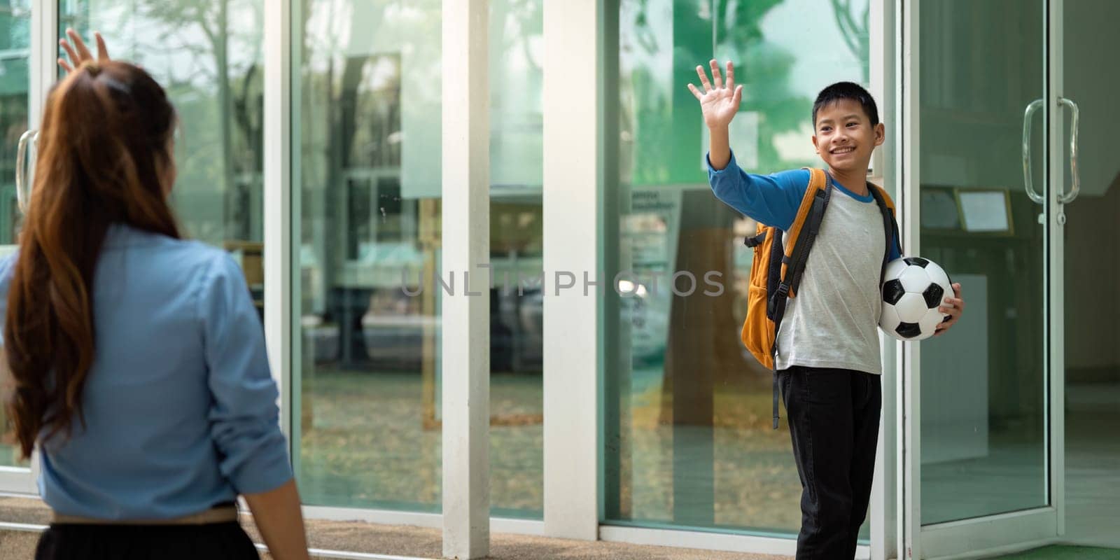 A cheerful boy waves goodbye to his mother, holding his soccer ball and backpack, signifying excitement and independence as he heads home from school.