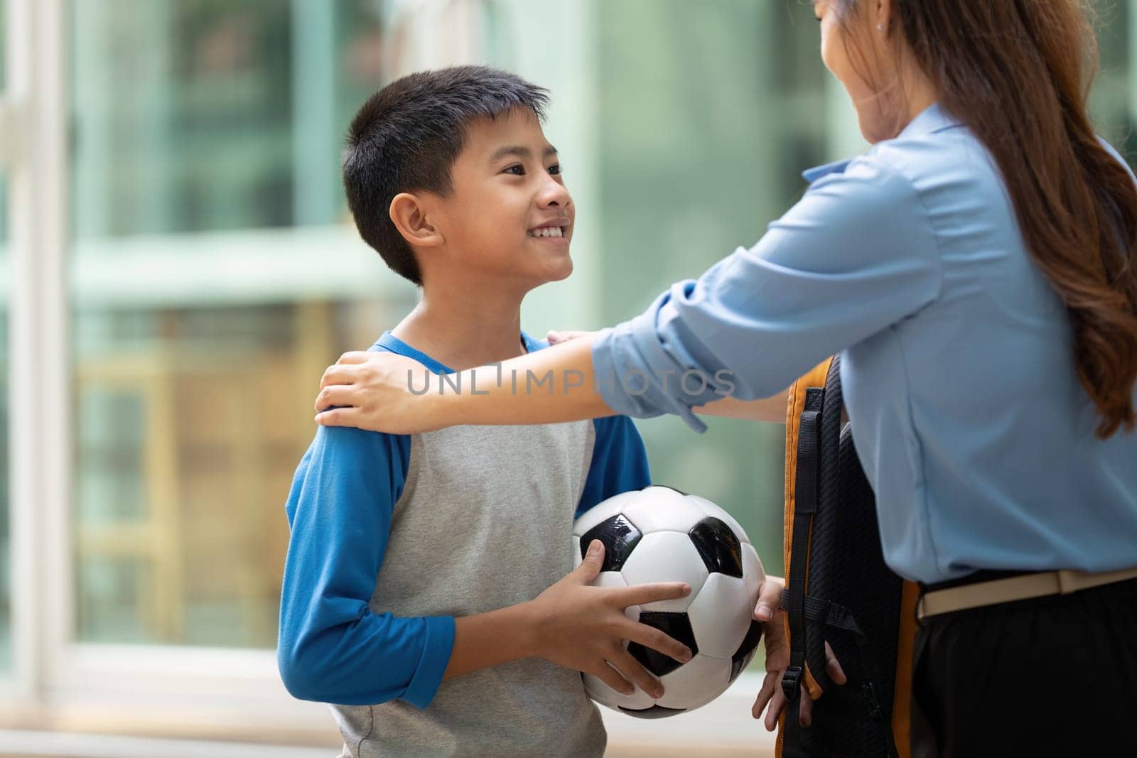 A warm interaction occurs in a school setting as a mother joyfully embraces her son after school, celebrating his accomplishments with a soccer ball in hand.