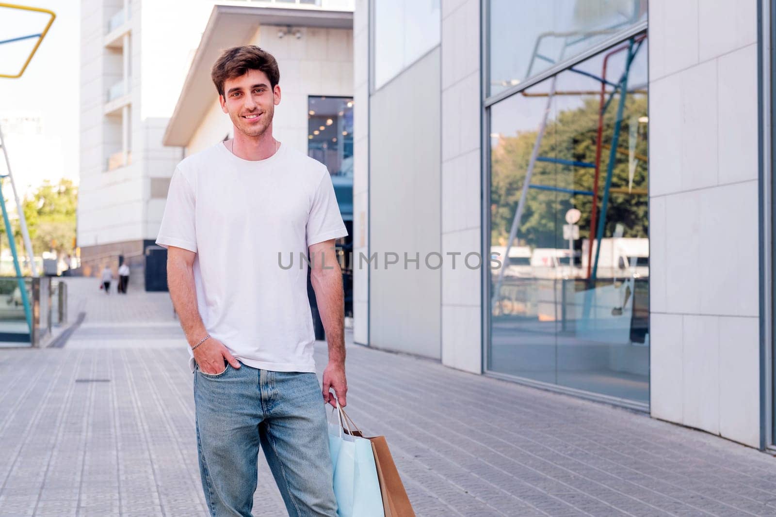 young adult man in casual clothes standing in a sidewalk near of a mall carrying shopping bags and smiling happy looking at camera, concept of youth and urban lifestyle, copyspace for text