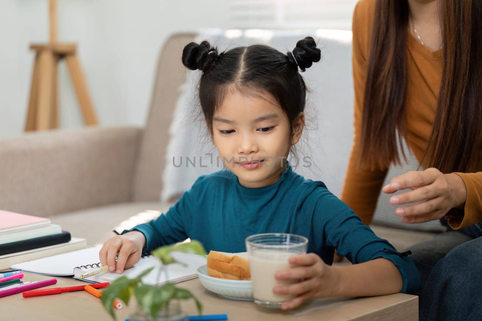 A young child sits at the table savoring a sandwich and milk while interacting with her mother, embracing the joyful moments of their morning routine before school.