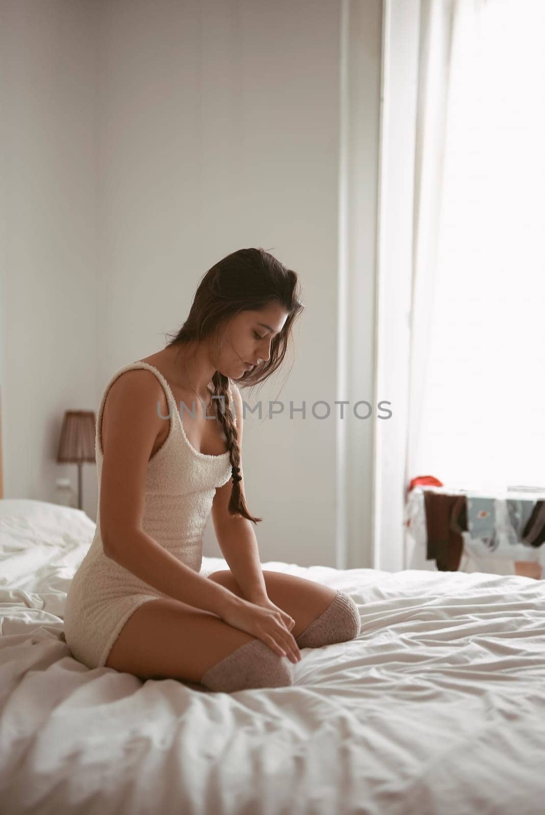 A young woman sits quietly and pensively on a neatly made bed, enveloped in the serene morning light