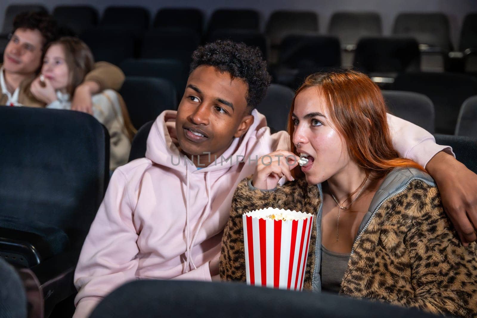 Young multi ethnic couple enjoying a movie and sharing popcorn in a cinema, surrounded by other people