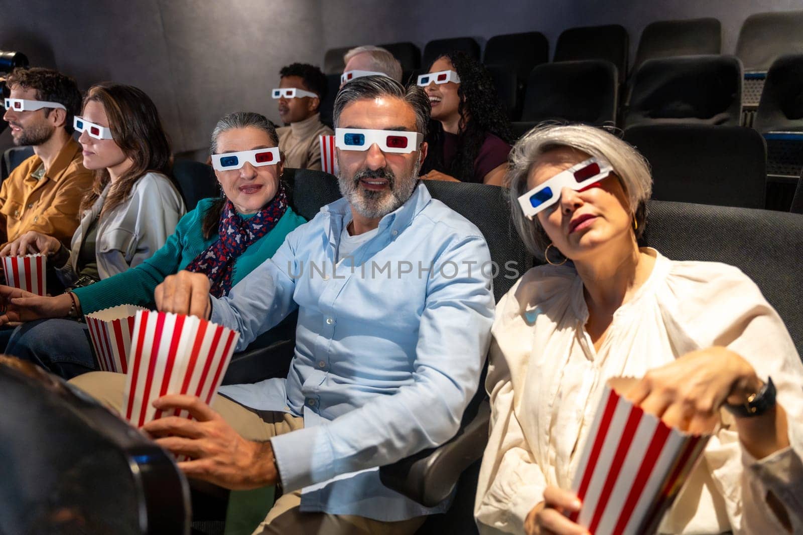 People of different ages and ethnicities wearing 3d glasses and eating popcorn while watching a movie in a cinema