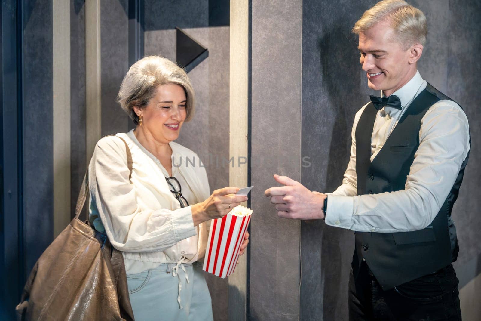 Elegant mature woman handing ticket to smiling usher while holding popcorn bucket, ready to enjoy movie session