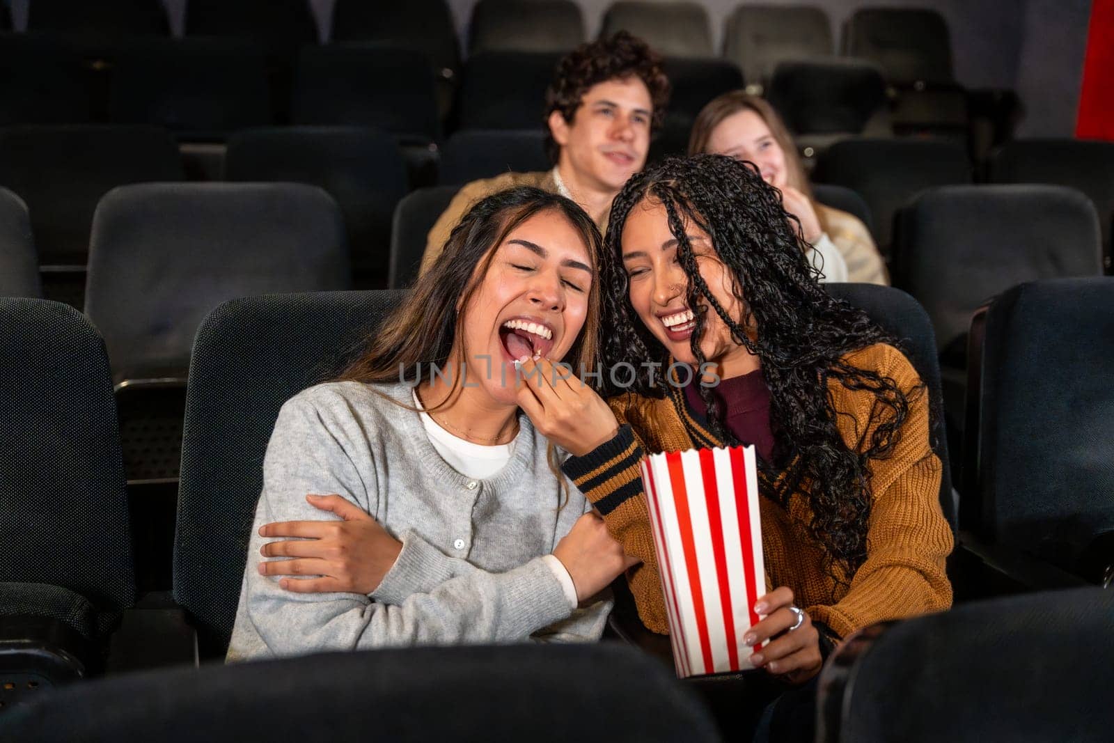 Two young women are enjoying a movie at the cinema, laughing and sharing popcorn, with other people in the background also watching the film
