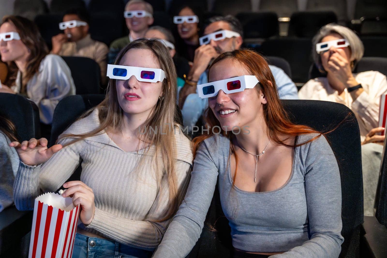 Two young women wearing 3d glasses are watching a movie and eating popcorn in a crowded movie theater