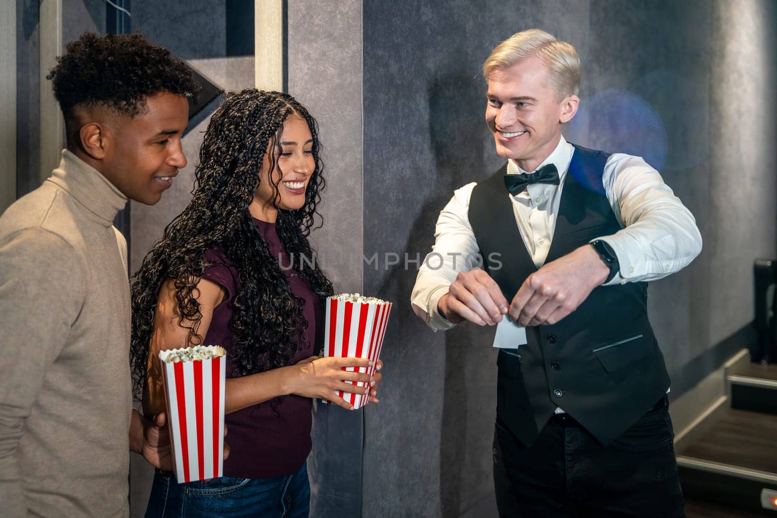 Young couple holding popcorn buckets entering a movie theater and checking their tickets with the usher before watching a film