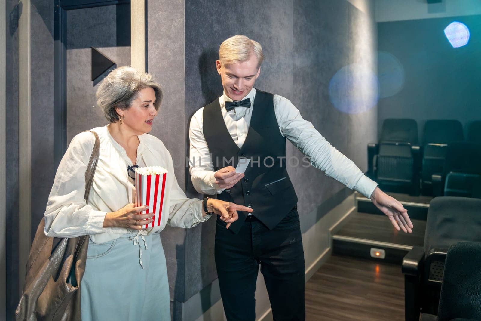 Cinema usher showing assigned seat to elegant mature woman holding popcorn bucket while entering movie theater