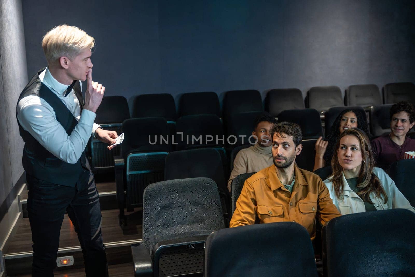 Young usher holding tickets and making silence gesture in movie theater while audience is watching film