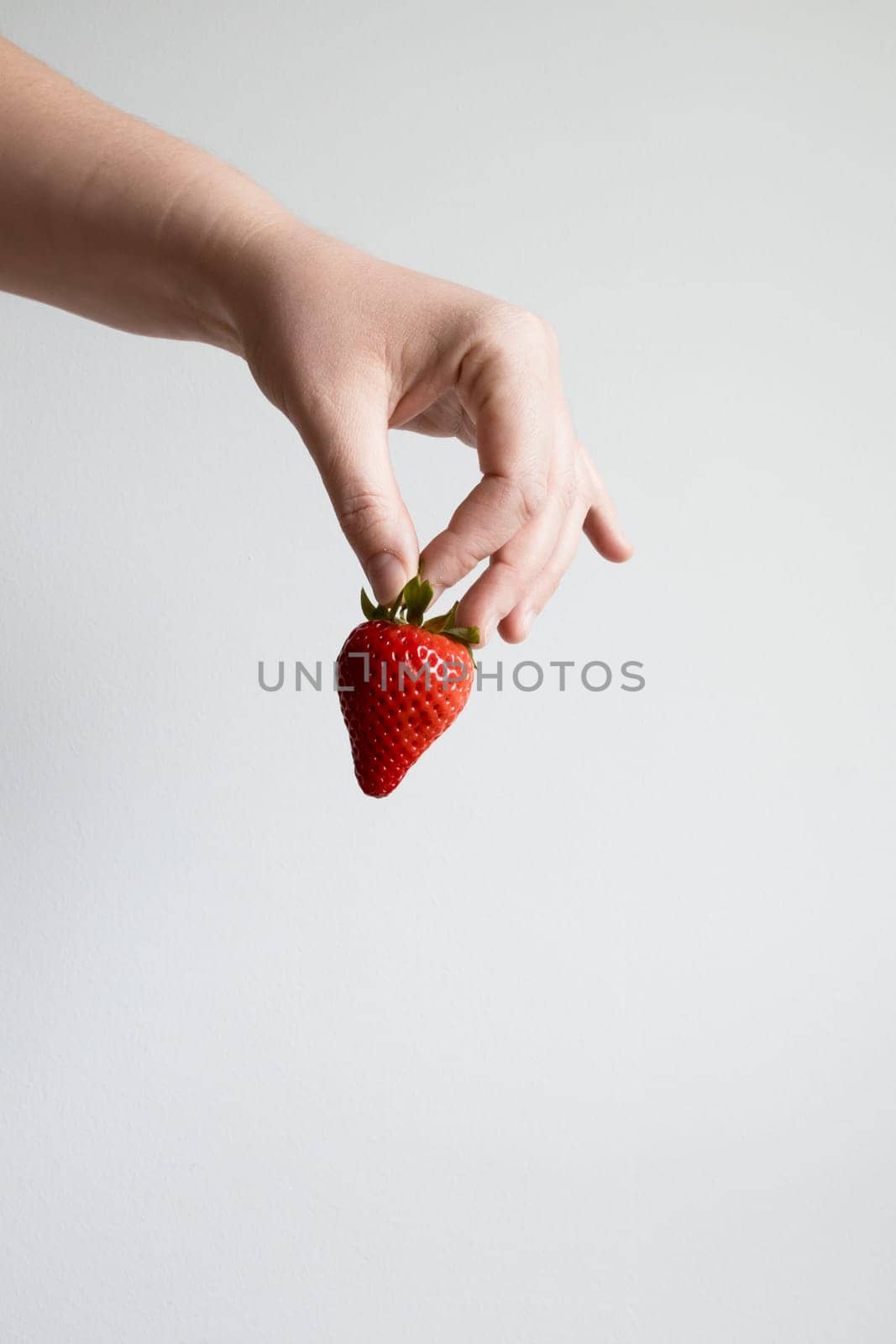 Hand holding fresh red strawberry against light grey background.