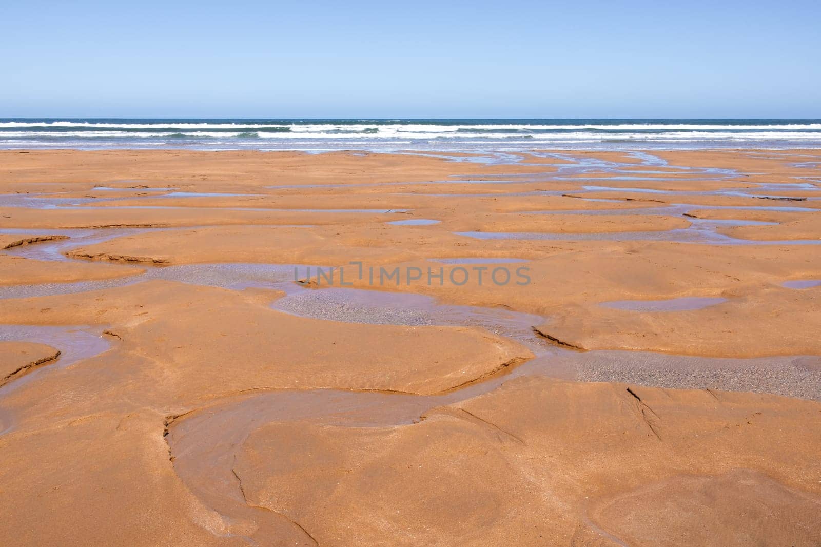 Vast sandy beach with puddles and ocean waves under clear blue sky.