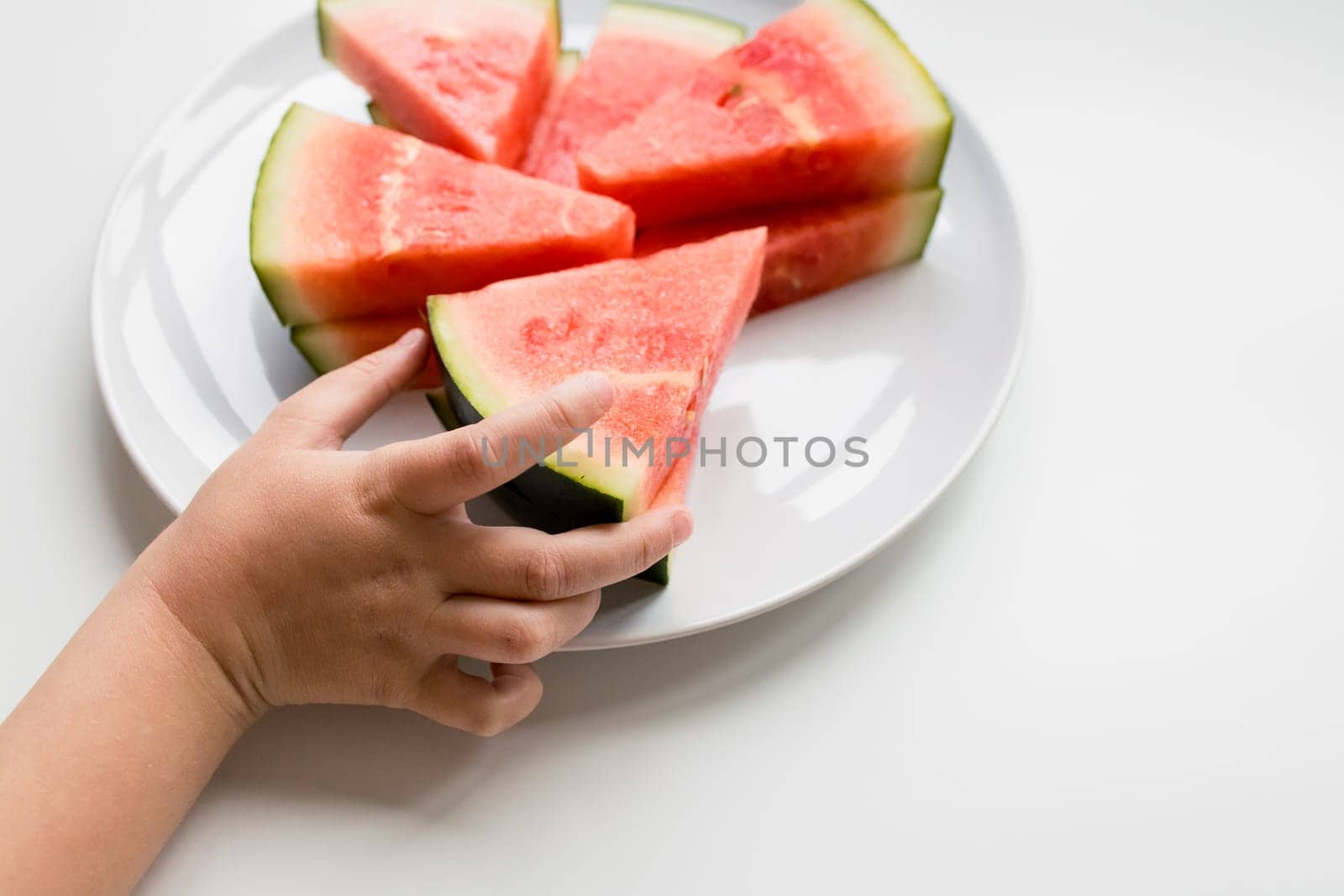 Child hand reaching for fresh watermelon slices on white plate.