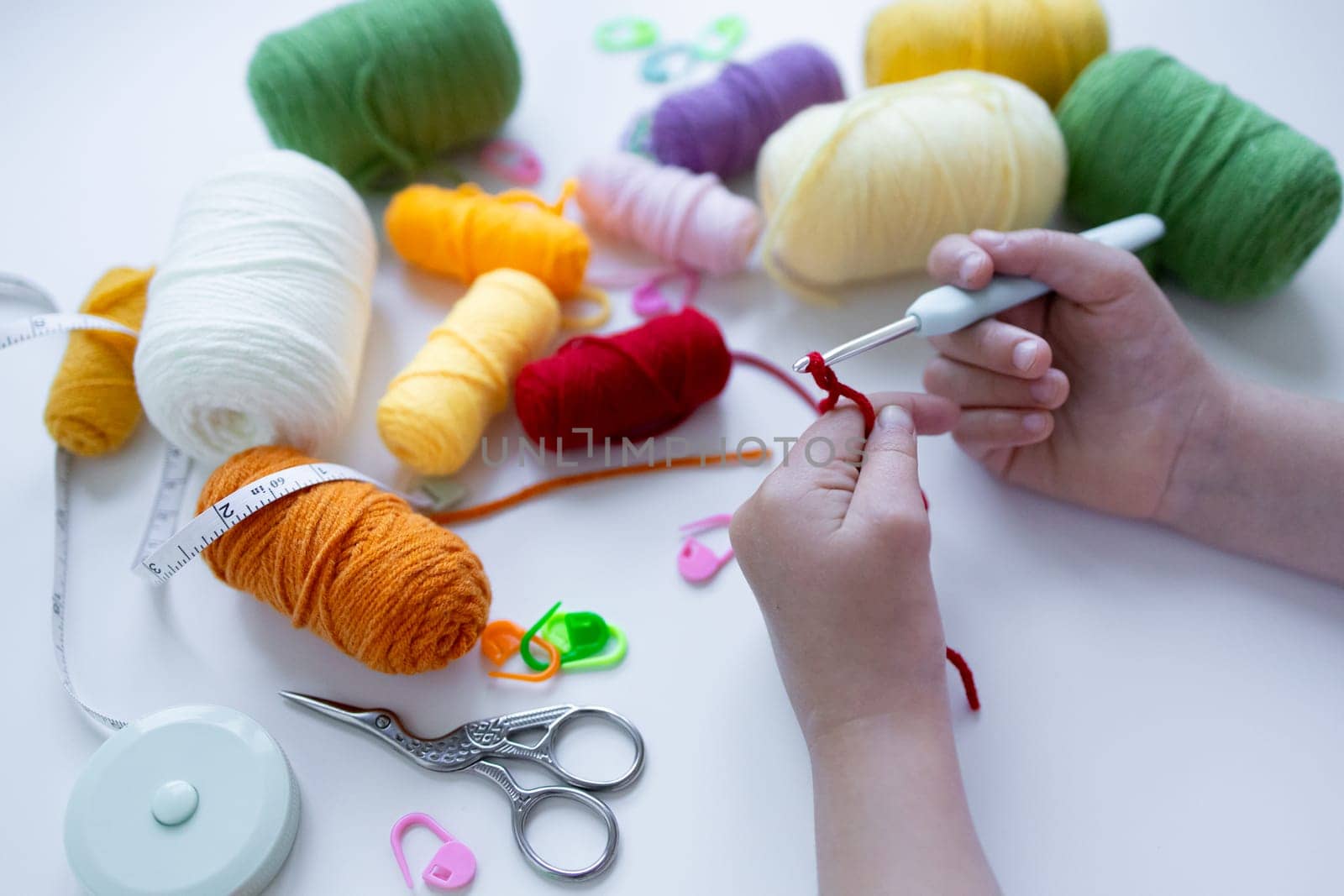 Child crocheting with colorful yarn and tools on white surface.