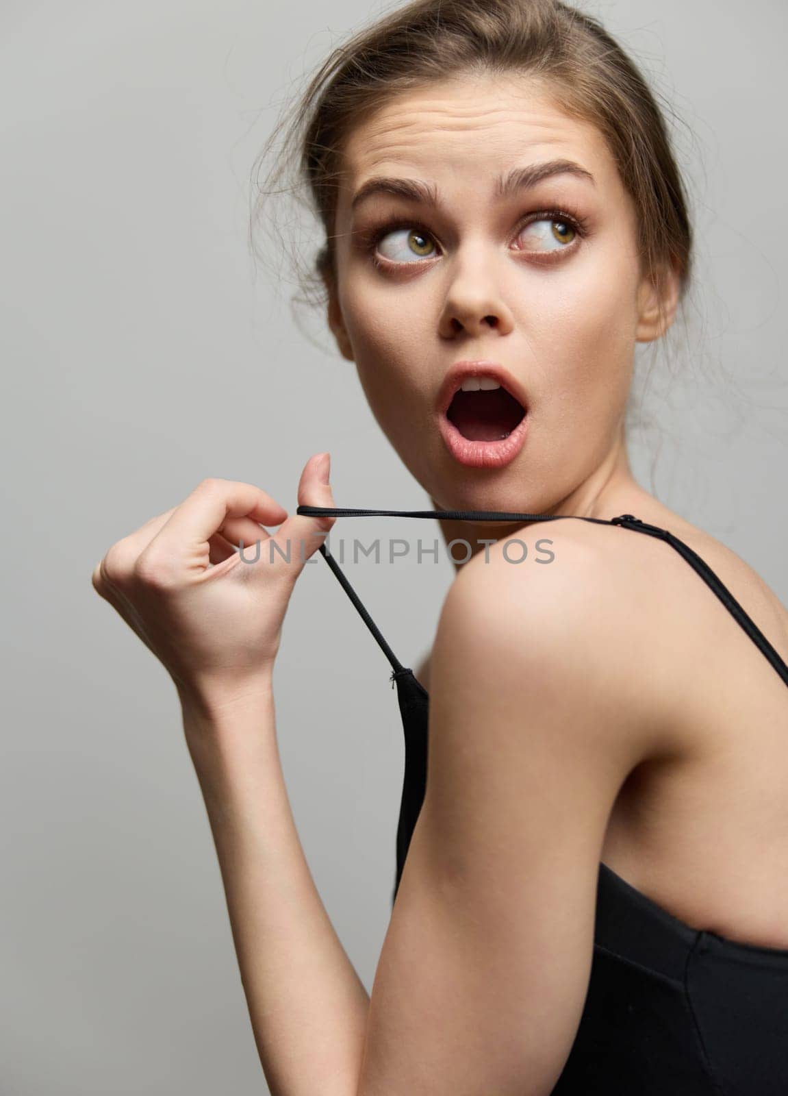 Expressive young woman with an astonished expression, touching her shoulder strap, creating a sense of intrigue and surprise in a minimalistic setting.