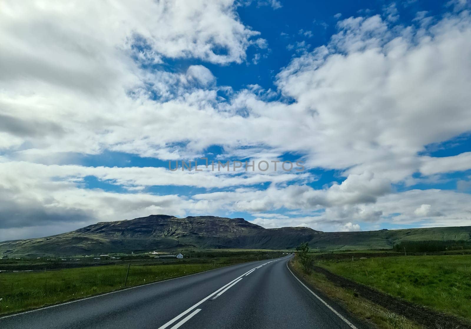 Asphalt road crossing green meadows with mountains and a cloudy sky in the background, typical Icelandic landscape