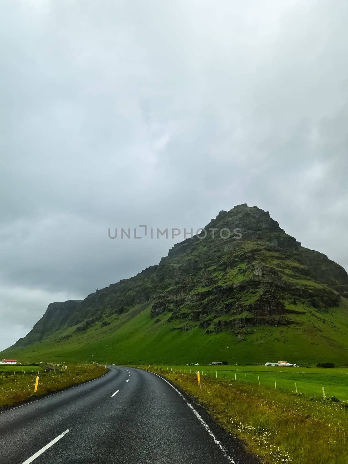Empty road leading towards a prominent green mountain under a cloudy sky in Iceland, showcasing the beauty of Icelandic nature